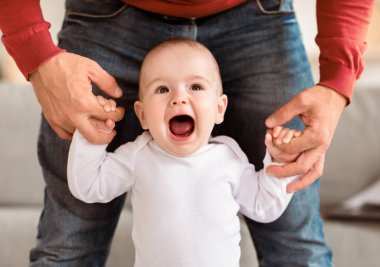 İlk Basamaklar. Mutlu küçük bebek evde babaların elini tutarak yürüyor. Baba küçük oğluna ilk adımını atması için yardım ediyor. Sevimli Aile Anları ve Babalık Konsepti. Kırpılmış