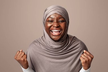 Başarı kutlaması yapan tesettürlü zenci Müslüman kadın kameraya karşı heyecanla el sıkışan yumruklar hafif arkaplanda poz veren duygusal Afrikalı İslamcı kadın, boş alan.