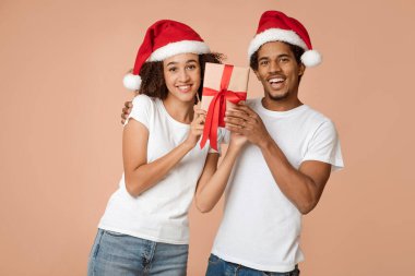 Birlikte Noel 'i kutluyoruz ve kameraya bakıyoruz. Milenyum Afrikalı Amerikalı bir adam Noel Baba şapkalı neşeli eşine Noel hediyesi veriyor, arka planda izole edilmiş, boş bir yer, stüdyo çekimi,