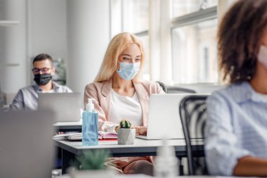 Ofisin içinde modern yönetici işyeri. Bin yıllık koruyucu maskeli sarışın kadın iş arkadaşlarıyla birlikte laptop 'da çalışıyor. Masadaki antiseptiğe odaklanıyor. Boş alan.