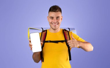 Çevrimiçi seyahat rezervasyon konsepti. Yakışıklı genç adam cep telefonunu boş ekranla gösteriyor ve mavi stüdyo arka planında el hareketi yapıyor. Web siteniz için boşluk