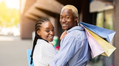Alışverişten sonra rahatla. Gülümseyen Afro-Amerikan çift, renkli çantalarıyla dükkandan eve yürüyorlar. Arkalarını dönüp kameraya, dışarı, fotokopi alanına bakıyorlar.