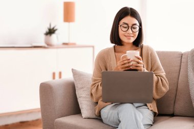 Kahve zamanı. Gözlüklü Asyalı bir kız öğrencinin portresi evde kahve içiyor ve bilgisayar kullanıyor, internette çalışıyor, kopyalama yapıyor. Gözlüklü kadın, ev ofisteki kanepede oturuyor.