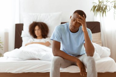 Ereksiyon bozukluğu, erkek gücü, kavga, seks sorunları ve ilişki. Üzgün, hayal kırıklığına uğramış, Afro-Amerikan bir erkek yatakta oturuyor, gücenmiş bir kadın yatak odasının parlak iç kısımlarına bakıyor, boş bir alan.