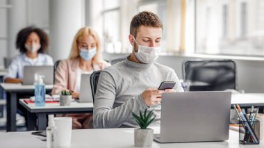 Karantina covid-19 sırasında çalışmak için modern teknoloji. İş yerinde koruyucu maskeli genç bir adam iş yerinde bilgisayar, bitki ve antiseptik ile akıllı telefonuyla iş arkadaşlarıyla yazı yazıyor.