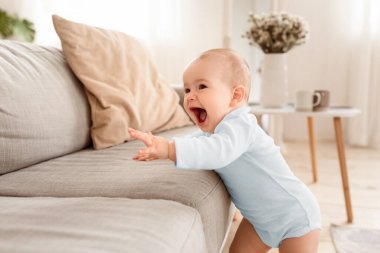 Evdeki oturma odasındaki kanepenin yanında gülüp eğlenen küçük kız. Sevimli küçük bebek ilk adımlarını yalnız atıyor. Mutlu Çocukluk ve Çocuk Bakımı Konsepti