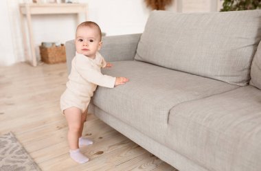Evdeki kanepenin yanında duran sevimli küçük bebek kameraya bakıyor. Tatlı çocuk oturma odasında ilk adımlarını yalnız atıyor. Çocukluk kavramı. Metin için Boş Boşluk