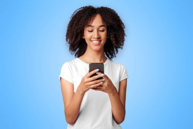 Mesaj ve modern blog. Gülümseyen bin yıllık Afro-Amerikan kadın pembe tişörtlü, kıvırcık saçlı, akıllı telefon tutan, yazan ve arayan, boş alan, stüdyo çekimi,