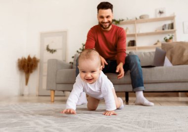 Mutlu genç baba, yeni yürümeye başlayan bebeğiyle oynuyor, babalık izni alıyor, küçük kızıyla evde bağ kuruyor. Çocuk Bakımı, Sevimli Babalık Anları, Ebeveynlik Mutluluğu. Seçici Odaklanma