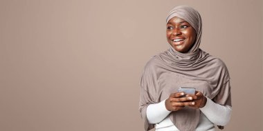 Mobil Teklif. Tesettürlü neşeli siyahi kadın akıllı telefon tutuyor ve fotokopi uzayına bakıyor, cep telefonu kullanarak ışık stüdyosunda duran gülümseyen kadın, modern iletişimin tadını çıkarıyor.