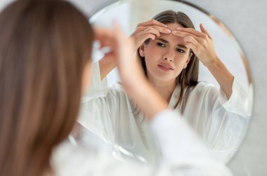 Problem Cildi. Banyoda aynanın yanında dikilen, suratını asan, acı çeken sivilceli, düşüncelere odaklanan genç bir kadın, yüzünü ekşiten bir kadın.