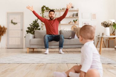 Komik baba evde koltukta oturan küçük bebek bebekle oynarken seçici bir şekilde babama odaklanıyor. Baba 'dan Çocuk' a. Dikkatini çekiyor. Babalık ve Babalık İzni