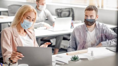 Coronavirüs salgını sırasında projeyle ilgili görüşmeler ve tartışmalar. Koruyucu maskeli genç adam ve kadın dizüstü bilgisayara bakar ve modern ofisin iş yerindeki cam levhalarla iletişim kurarlar.