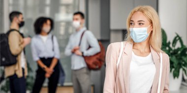 Tecritten sonra işe dön ve yeni normal haline dön. İş elbisesi giymiş koruyucu maskeli bir kadın modern ofisin koridorunda bulanık iş arkadaşlarıyla boş alana bakıyor.