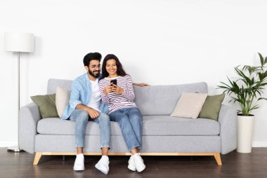 Rahat, neşeli, genç, çok kültürlü çift orta doğulu erkek ve Hintli kadın evdeki koltukta oturuyor, akıllı telefon kullanıyor, gülümsüyor, internetten alışveriş yapıyor, internette sörf yapıyor, uzay kopyaları yapıyor.