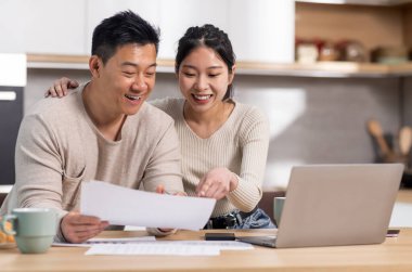 Cheerful asian spouses paying bills on Internet from home, sitting together at kitchen table, holding letters and papers, using computer and smiling, enjoying easy e-banking, copy space