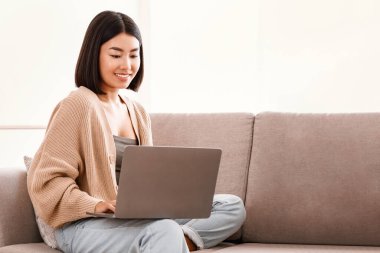 Teknoloji. Milenyum Asyalı Kadın dizüstü bilgisayarla evde çalışıyor, serbest çalışmanın keyfini çıkarıyor, koltuğunda bilgisayarla oturan, klavyede yazan, video izleyen ya da öğrenen genç bir kadın.