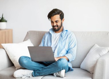 Uzak çalışma. Mutlu Arap Serbest Çalışan Adam evde dizüstü bilgisayar kullanıyor, klavyede daktilo kullanıyor, genç doğu çocuğu oturma odasında oturuyor, uzaktan internet projesi üzerinde çalışıyor, Boşluğu Kopyala