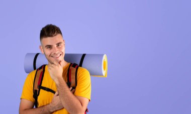 Sırt çantası ve turist minderiyle gülümseyen ve mavi stüdyo arka planında çenesine dokunan yakışıklı adamın portresi. Seyahat, turizm, kamp, yürüyüş konsepti