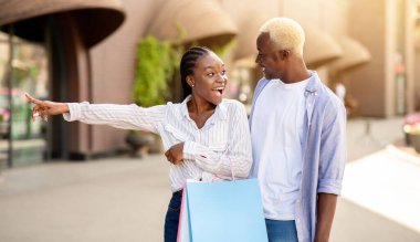 Marka etiketleri ve özel indirimler. Şaşkın Afro-Amerikalı kız alışveriş çantaları ile sokakta erkeklere gösteri yapıyor.
