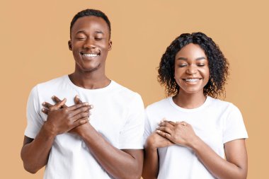 Sarı stüdyo arka planında gözleri kapalı dua eden siyah çiftin yakın çekimi. Kutsanmış Afrikalı Amerikalı erkek ve kadın avuçlarını göğsünde tutuyor ve gülümsüyor, en iyisini umut ediyor.