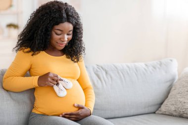 Mutlu Afro-Amerikalı, bebek ayakkabılarıyla oynayan bir kadın bekliyor. Karnında bebek ayakkabıları taşıyan hamile bir zenci kadın. Çocuğunu bekliyor. Evdeki kanepede dinleniyor.