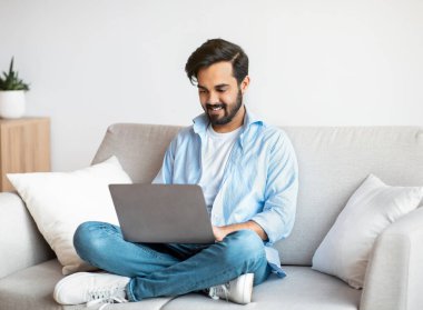 Uzak çalışma. Mutlu Arap Serbest Çalışan Adam evde dizüstü bilgisayar kullanıyor, klavyede daktilo kullanıyor, genç doğu çocuğu oturma odasında oturuyor, uzaktan internet projesi üzerinde çalışıyor, Boşluğu Kopyala