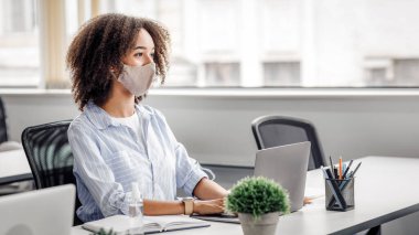 Çalışmak için ortak çalışma yeri. Koruyucu maskeli Afrikalı Amerikalı bir kadın müşterisine ya da meslektaşına bakar ve dizüstü bilgisayarında çalışır. Bilgisayarlı ve esrar dolu bir masada oturur. Virüs salgını sırasında, boş bir alanda.