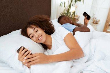 Covid-19 tecridi sırasında sosyal ağlar, cihazlar ve çevrimiçi iletişim. Gülümseyen Afro-Amerikan milenyum bayanı akıllı telefona bakıyor ve rahat yatak odasında kocasından uzaklaşıyor.