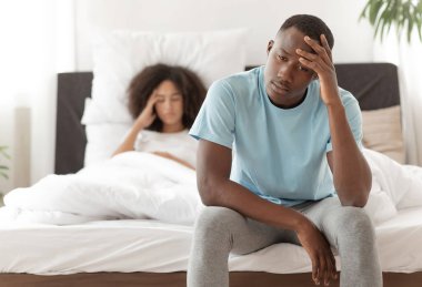 Baş ağrısı, iktidarla ilgili sorunlar, erkek iktidarı, kavga ve skandallar. Milenyum Afrikalı Amerikalı adam yatağa oturup elini alnına bastırıyor modern yatak odasında üzgün bir eş, fotokopi çekiyor.