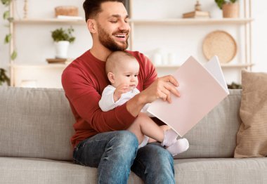 Genç baba, küçük kız bebek için kitap okuyor. Evde yeni yürümeye başlayan çocukla oturuyor. Babacığın Babalığın tadını çıkarıyor. Babalar Sevgi ve Sevimli Ebeveynlik Anları