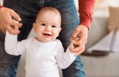 Gülümseyen bebek ilk adımı atıyor. Babaların elini tutuyor. Evde yürüyor. Sevgi dolu bir baba, küçük kızının içeri girmesine yardım ediyor. Sevimli Babalık Anları Konsepti. Kırpılmış