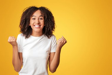 Mutluluk ve başarı duyguları. Kıvırcık saçlı, gülümseyen genç Afro-Amerikan kadını. Yumruklarını kaldır ve çevrimiçi zafere sevin. Sarı arka planda izole edilmiş, boş alan, stüdyo çekimi.