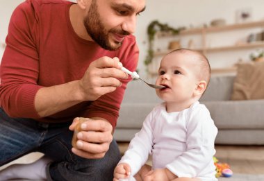 Sevgi dolu genç baba Spoon 'ın bebeğini beslemesi bebek bakımı sırasında oturma odasında yerde oturması. Küçük Çocuk İçin Sağlıklı Beslenme. Kırpılmış