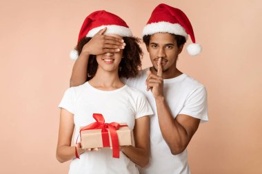 Yeni yıl hediyeleri, kış tatilleri. Şapkalı Afrikalı Amerikalı bir adam sessizlik işareti gösteriyor, karısına gözlerini kapıyor ve ona ışık arka planda izole edilmiş bir hediye kutusu veriyor, stüdyo çekimi, boş alan.