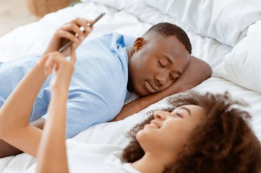 Çevrimiçi sohbet, teknoloji ve evde Coronavirus sırasında ilişkiler. Bin yıllık gülümseyen Afro-Amerikan kadın akıllı telefonda sörf yapıyor, erkek yatak odasındaki yumuşak beyaz yatakta uyuyor, içi boş.