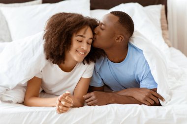 Aşk, ilişkiler, nazik, aile ve romantizm. Mutlu, genç, yakışıklı Afrikalı, Amerikalı koca, kadını alnından öpüyor, beyaz örtülerin altında yatıyor, rahat yatak odasında sessizliğin tadını çıkarıyor, özgür bir yerde.