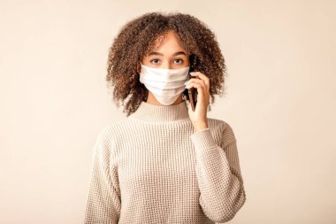Virüs salgını sırasında karantina sonrası sosyal bağlantı ve mesafe. Koruyucu maskeli Afrikalı Amerikalı kadın telefonda konuşuyor. Açık krem arka planda izole edilmiş, boş alan, stüdyo çekimi.