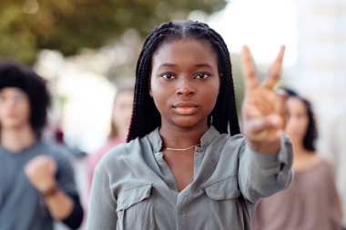 Uluslararası protestocuları yöneten genç siyahi kadın, kameraya barış jesti gösteriyor. Dünya barışı için savaşan, sokaklarda yürüyen çok ırklı göstericiler.
