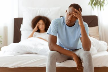 Baş ağrısı, iktidarla ilgili sorunlar, erkek iktidarı, kavga ve skandallar. Milenyum Afrikalı Amerikalı adam yatağa oturup elini alnına bastırıyor modern yatak odasında üzgün bir eş, fotokopi çekiyor.