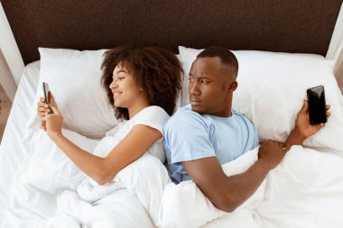 Aldatma, aldatma, şüphe, teknoloji ve ilişki sorunları. Gerçekten ilgilenen bin yıllık Afro-Amerikan erkeği, yatak odasında gülümseyen karısının akıllı telefonuna bakıyor.