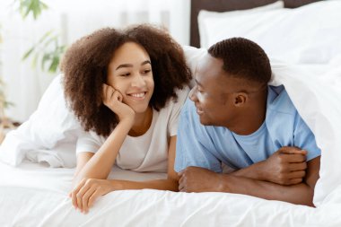 Genç aile rahatlar, romantik sohbet eder ve hafta sonu evde boş vakit geçirir. Yorgan altında mutlu, genç Afrikalı Amerikalı çift yatakta konuşuyor, birbirlerine bakıyor ve yatak odasında dinleniyorlar.