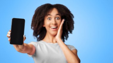 Nakit para, online galibiyet ve inanılmaz bir mesaj. Şaşırmış, mutlu, Afro-Amerikan kadın, günlük giysiler içinde, yüz yüze ütülüyor ve menekşe arka planında boş ekranlı akıllı telefon gösteriyor.