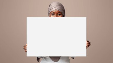Reklam için Alan Kopyala. Tesettürlü Siyah Kadın Boş Beyaz Plaketin Arkasına Saklanıyor, Yazı veya Tasarım İçin Boş Yer Gösteriyor, Müslüman Kadın Işık Stüdyosu Arkaplanının Başında Duruyor