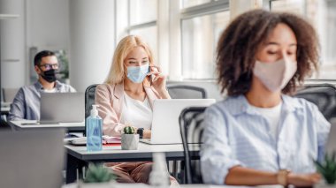 Şirket çalışanları ofiste çalışıyor. İş yerlerindeki koruyucu maskeli, antiseptik ve dizüstü bilgisayarlı çok ırklı yöneticiler, sarışına odaklan, koronavirüs salgını sırasında telefonla konuş, boşluğu kopyala