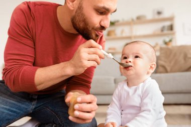 Evde yerde oturan kaşıkla oğlunu besleyen genç bir baba. Babam çocuk bakıcılığı yapıyor. Babalık izni, Babalık, Yeni Doğan Bebekler İçin Sağlıklı Yemek ve Beslenme