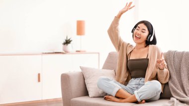 Boş zaman kavramı. Aşırı mutlu Asyalı kadın portresi. Kablosuz kulaklık takıyor, en sevdiği şarkıyı dinliyor oturma odasında kanepede oturuyor. Milenyum bayanı gözleri kapalı dans ediyor.