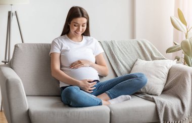 Evdeki kanepede dinlenirken karnını kucaklayan hamile bir kadın. Oturma odasındaki koltukta dinlenen hamile bir kadın. Hamileliğinin ve gelecekteki hamileliğinin tadını çıkarıyor.