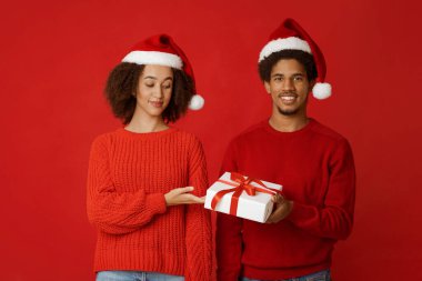 Kış bayramı kutlaması. Genç Afrikalı Amerikalı gülümseyen bir adam Noel hediyesi sunuyor, elinde hediye paketi var, ilgilenen bir kadın var, kırmızı arka planda izole edilmiş, stüdyo fotoğrafı.