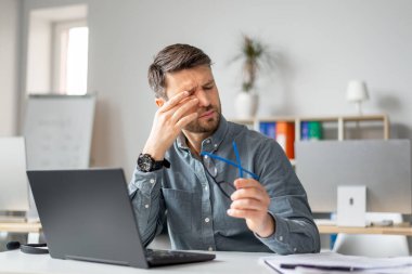 Yorgun, olgun bir adam, göz yorgunluğu çekiyor, gözleri ovuşturuyor, bilgisayar işinden yorgun düşmüş, baş ağrısı çekiyor, ofiste otururken görme sorunu yaşıyor.